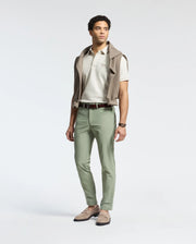 A man stands against a plain background, dressed in a white polo shirt and Psycho Bunny's MENS MAVERICK 10K STEP PANT - B6P788D200. The five-pocket styling and light green, four-way stretch fabric enhance his casual yet polished look, completed with loafers and a sweater draped over his shoulders.