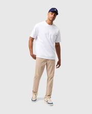 A man stands against a plain background wearing a white T-shirt, Psycho Bunny's MENS MAVERICK 10K PANT (B6P788D200), blue-and-white sneakers, and a navy blue cap. He has one hand in his pocket and a relaxed expression on his face.