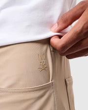 Close-up of a person wearing beige MENS MAVERICK 10K PANTS - B6P788D200 by Psycho Bunny, featuring a small, embroidered logo of a rabbit skull and crossbones above the front pocket. The person is also wearing a white shirt tucked in and is slightly lifting it to reveal the logo.