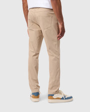 A person is wearing Psycho Bunny MENS MAVERICK 10K PANT - B6P788D200 in light khaki with back pockets. They have on white and blue sneakers with tan accents. The person is standing, showing the back view of their legs and a portion of a white shirt. The background is plain and light-colored.