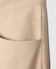 Close-up of a beige Psycho Bunny MENS MAVERICK 10K PANT - B6P788D200 pocket featuring an embroidered logo of a rabbit with crossbones underneath. The stitching and texture of the fabric, along with its four-way stretch, are clearly visible, emphasizing the detailed design and craftsmanship.