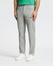 A man wearing Psycho Bunny MENS MAVERICK 10K STEP PANT - B6P788D200 in light gray, styled with five pockets, a green shirt, and white sneakers stands with one hand in his pocket against a plain white background.