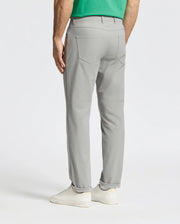 A man faces away wearing light gray Psycho Bunny MENS MAVERICK 10K STEP PANT - B6P788D200, featuring five-pocket styling, paired with a green shirt and white sneakers, against a plain white background.