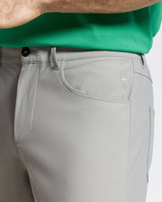A person wears Psycho Bunny’s MENS MAVERICK 10K STEP PANT - B6P788D200 in light gray, featuring four-way stretch, five-pocket styling, and a bunny and crossbones logo on the front pocket, paired with a bright green shirt.