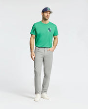 A man wears a green Psycho Bunny tee, white sneakers, a blue and white cap, and the MENS MAVERICK 10K STEP PANT - B6P788D200 featuring five-pocket styling and four-way stretch, standing against a plain white background.