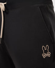 Close-up of the upper section of a pair of MENS JAMES PREMIUM SWEATPANT - B6P791D200 by Psycho Bunny, showcasing black sweatpants with an elastic waistband and beige drawstrings. Crafted from a soft cotton-blend for ultimate comfort, these premium bottoms feature an embroidered beige logo of a bunny head with crossbones just below the front right pocket.