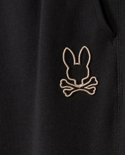 Close-up image of black, soft cotton-blend fabric on the MENS JAMES PREMIUM SWEATPANT - B6P791D200 by Psycho Bunny, featuring an embroidered design of a bunny head above two crossed bones, stitched in light-colored thread.