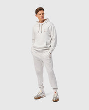 A person dons the MENS JAMES PREMIUM SWEATPANT in light gray cotton-blend by Psycho Bunny. They stand with hands in pockets, sporting an outfit that includes a subtle embroidered design. Complementing this look are sneakers with gray and beige accents on a plain background, highlighting ultimate comfort.