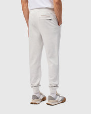 A person dressed in a white T-shirt is sporting the MENS JAMES PREMIUM SWEATPANT - B6P791D200 by Psycho Bunny, made from a comfortable cotton blend in light gray. With hands tucked into the pockets, the sweatpants highlight a subtle "Psycho Bunny" logo on the back and are combined with gray and white sneakers featuring brown soles against a simple background.