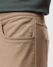 Close-up of the MENS HAYS TWILL PANT by Psycho Bunny, crafted from a cotton-blend twill in a tan color with a pocket featuring an embroidered bunny logo. The pants are designed for ultimate comfort and are worn with a dark-colored shirt tucked in, highlighting the garment's intricate stitching details.