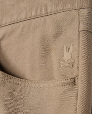 Close-up of the tan twill fabric from Psycho Bunny's MENS HAYS TWILL PANT - B6P894D200, featuring a back pocket adorned with an embroidered logo of a rabbit's head above crossed bones. The detailed stitching enhances the texture, ensuring ultimate comfort.