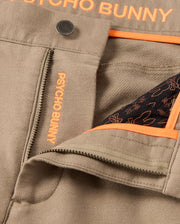 Close-up of the MENS HAYS TWILL PANT - B6P894D200 in a beige cotton-blend twill, featuring an open zipper that reveals interior black fabric adorned with orange patterns. The brand name "Psycho Bunny" stands out on the zipper placket and waistband, ensuring ultimate comfort and style with each wear.