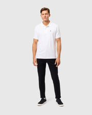 Against a plain white backdrop, a man stands wearing a white polo shirt paired with Psycho Bunny's Men's Hays Twill Pant (B6P894D200) in black, crafted from cotton-blend twill for ultimate comfort. His look is completed by black sneakers, and his short brown hair and neutral expression enhance the clean, relaxed style.