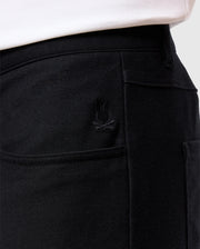 A close-up of a person wearing Psycho Bunny's MENS HAYS TWILL PANT - B6P894D200 reveals the ultimate comfort and style of these black cotton-blend twill pants. Highlighted by subtle rabbit embroidery on the side, they are paired with a white top to emphasize their texture and design, featuring 4-way stretch for optimal mobility.