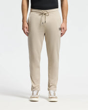 A person wearing Psycho Bunny's beige MENS MONROE TWILL PATCH FLEECE SWEATPANT featuring a drawstring and small embroidered logo on the left pocket, paired with a black shirt and white sneakers, stands against a plain white background.