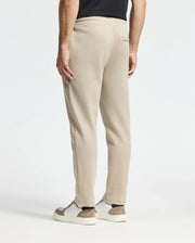 A person is seen from behind wearing beige Monroe twill patch fleece sweatpants by Psycho Bunny and a black shirt. The look is complemented by white sneakers with brown accents against a plain white background, emphasizing the casual outfit.