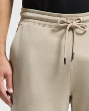 Close-up of an individual wearing Psycho Bunny's MENS MONROE TWILL PATCH FLEECE SWEATPANT in beige, featuring a drawstring and side pockets. They pair it with a black top, with their left hand partially visible at their side.