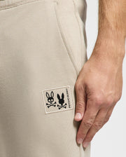A close-up highlights a hand resting on the MENS MONROE TWILL PATCH FLEECE SWEATPANT - B6P929D200 by Psycho Bunny, focusing on the patch displaying a bunny head above crossed bones.