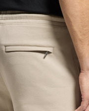 A close-up shows a person facing away, wearing Psycho Bunny's beige Monroe Twill Patch Fleece Sweatpants (B6P929D200). A small silver pin is partially in the back pocket, with part of their right arm visible against a dark background.
