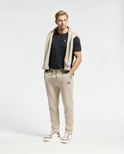 A man in a black T-shirt with a light beige sweater draped over his shoulders, wearing Psycho Bunny's Monroe Twill Patch Fleece Sweatpants and white sneakers, stands against a plain white background.