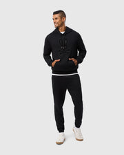 A person confidently stands with hands in pockets, wearing a black hoodie featuring a chenille bunny skull logo patch. The look is completed with Psycho Bunny's MENS PACIFIC CHENILLE JOGGER - B6P972D200 and white sneakers, all set against a light gray background.