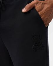 Close-up of a person wearing the Psycho Bunny MENS PACIFIC CHENILLE JOGGER - B6P972D200 with their hand in the pocket. The joggers feature an embroidered skull and crossbones design with bunny ears on the side, complemented by a chenille logo patch that adds a unique touch to this casual bottom, set against a plain white background.