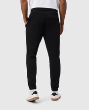 A person wearing Psycho Bunny's MENS PACIFIC CHENILLE JOGGER - B6P972D200, which features a chenille logo patch, along with white sneakers, stands with their back to the camera against a plain gray background.