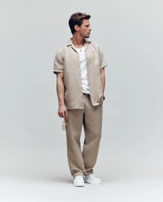 A man wears the Psycho Bunny MENS WINDCREST LINEN BLEND SHORT SLEEVE SHIRT (B6Q148C200) in beige over a white tee, with matching beige pants and white sneakers. He holds sunglasses and looks left, set against a plain light background.