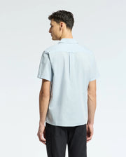 A man stands with his back to the camera in black pants and the Psycho Bunny MENS COBAR WOVEN SHIRT - B6Q522F200, against a plain light background.