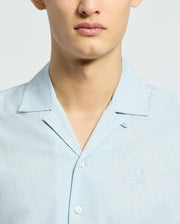 Close-up of a person in the Psycho Bunny MENS COBAR WOVEN SHIRT - B6Q522F200, a light blue linen-blend short sleeve shirt with collar and tonal embroidery on the left chest. Only the lower part of their face is visible.