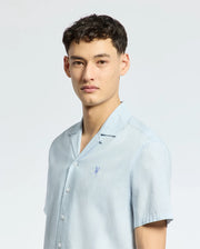 A young man with short dark hair wears a Psycho Bunny MENS COBAR WOVEN SHIRT (B6Q522F200) in light blue, standing against a plain light background and looking slightly to the side with a neutral expression.