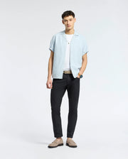 A man poses against a plain background wearing the Psycho Bunny MENS COBAR WOVEN SHIRT - B6Q522F200 over a white tee, with black pants, beige loafers, a belt, and a wristwatch. He faces the camera in a relaxed stance.
