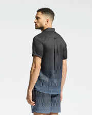 A man models the Psycho Bunny MENS BENDIGO TENCEL SHIRT - B6Q627F200, a short-sleeved camp collar shirt paired with matching shorts, featuring a black and blue geometric pattern against a plain background.