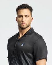 A person with short curly hair wears the MENS BENDIGO TENCEL SHIRT by Psycho Bunny, a black short-sleeved shirt featuring a small blue logo on the chest and a subtle ombre effect. The background is plain white.