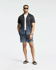 A man in Psycho Bunny's MENS BENDIGO TENCEL SHIRT, featuring a black ombre design and camp collar, stands confidently in matching shorts. A white T-shirt underneath and black slide sandals polish off his look against a plain white backdrop.