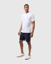 A man stands against a plain light gray background, wearing a white polo shirt and Psycho Bunny MENS WILLIS STRETCH TENCEL SHORT - B6R239Y1WB in dark navy, paired with white sneakers featuring brown soles. He has a relaxed and confident pose, looking straight ahead.