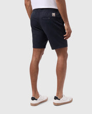 A person is shown from the back, wearing a white t-shirt, Psycho Bunny MENS WILLIS STRETCH TENCEL SHORT - B6R239Y1WB with a small rectangular patch and a single back pocket, and white sneakers with black accents. The background is plain and light gray.