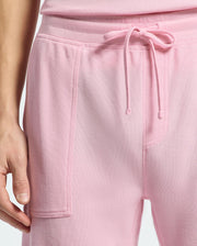 A person is wearing Psycho Bunny's MENS TAYLOR LIGHT PIQUE SWEATSHORT in light pink, featuring a drawstring waist and a front pocket. Their left hand rests near the pocket, completing the casual-chic look against a light gray background.