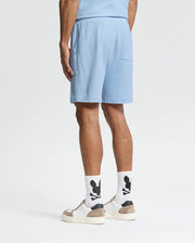 A person faces away, wearing Psycho Bunny's MENS TAYLOR LIGHT PIQUE SWEATSHORT (B6R344F200) in light blue and a coordinating top. They have white socks with a black pattern and white sneakers with beige soles against a plain white backdrop.