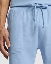 Close-up of someone wearing Psycho Bunny's MENS TAYLOR LIGHT PIQUE SWEATSHORTS, with a drawstring and side pocket. A hand is visible, accentuating the light piqué fabric's texture.
