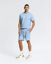 A person stands against a plain background in a casual-chic outfit, wearing a light blue T-shirt, Psycho Bunny MENS TAYLOR LIGHT PIQUE SWEATSHORTS, white socks, and white sneakers with beige accents. With short hair and their left hand in their pocket, they blend comfort with style.