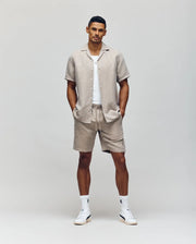 A man faces forward against a plain background, wearing a white tee, light beige short-sleeve button-up, Psycho Bunny MENS WINDCREST LINEN BLEND DRAWSTRING SHORT - B6R474C200, white socks, and sneakers. His hands rest in his pockets.