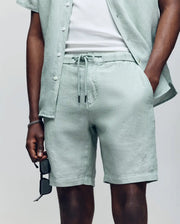 Wearing Psycho Bunny’s MENS WINDCREST LINEN BLEND DRAWSTRING SHORT (B6R474C200) and a matching light green shirt over a white T-shirt, the model poses with one hand in a pocket and black sunglasses in the other; face not shown.