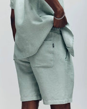 A person stands with one hand in the pocket of Psycho Bunny's MENS WINDCREST LINEN BLEND DRAWSTRING SHORT (B6R474C200), paired with a matching light green shirt. The image shows only their lower torso and upper legs against a plain background.
