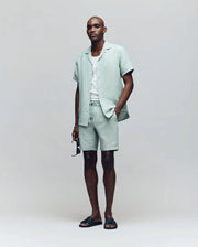 A man stands against a light gray background wearing Psycho Bunny’s MENS WINDCREST LINEN BLEND DRAWSTRING SHORT (B6R474C200), a pale green short-sleeve button-up shirt, white T-shirt, black slide sandals, and holding sunglasses.