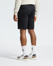 A person is wearing a cream long-sleeve shirt, Psycho Bunny's Levanzo Embroidered Micro French Terry Sweatshorts, and white socks with black logos, paired with white and gray sneakers. The Embroidery Bunny on the shorts adds charm as they stand against a plain background, shown from behind.