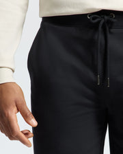 A person is wearing Psycho Bunny's black Levanzo embroidered micro French terry drawstring pants paired with a white long-sleeve shirt. One relaxed hand is visible by their side, focusing on the comfort and drawstring detail reminiscent of the Levanzo Sweatshort style.