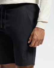 Wearing Psycho Bunny's black Levanzo Embroidered Micro French Terry Sweatshorts with a rabbit and crossbones logo near the hem, the person pairs it with a white long-sleeve shirt, while their right hand rests beside these comfort-focused shorts featuring a handy drawstring.