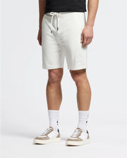 A person in Psycho Bunny MENS LEVANZO EMBROIDERED MICRO FRENCH TERRY SWEATSHORT (B6R578F200), a black t-shirt, white crew socks with black designs, and white sneakers with beige accents stands against a plain background.