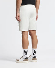 A person seen from behind wears a black shirt, Psycho Bunny Mens Levanzo Embroidered Micro French Terry Sweatshorts (B6R578F200) in white, white socks with black graphic, and white sneakers with gray details, against a plain white background.
